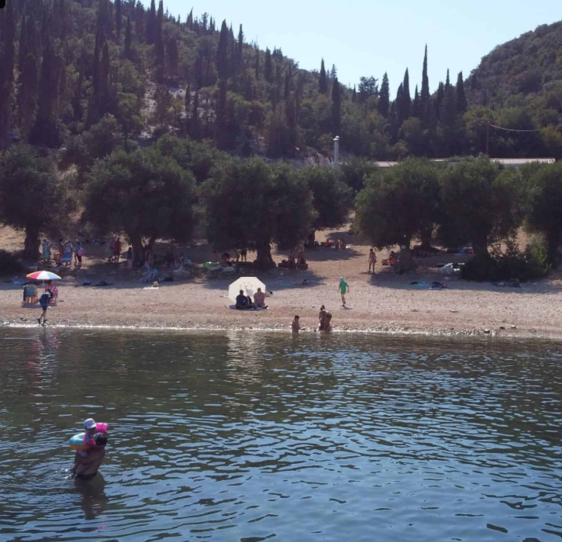 Foki Beach scene with people, trees, and calm water in Fiskardo.